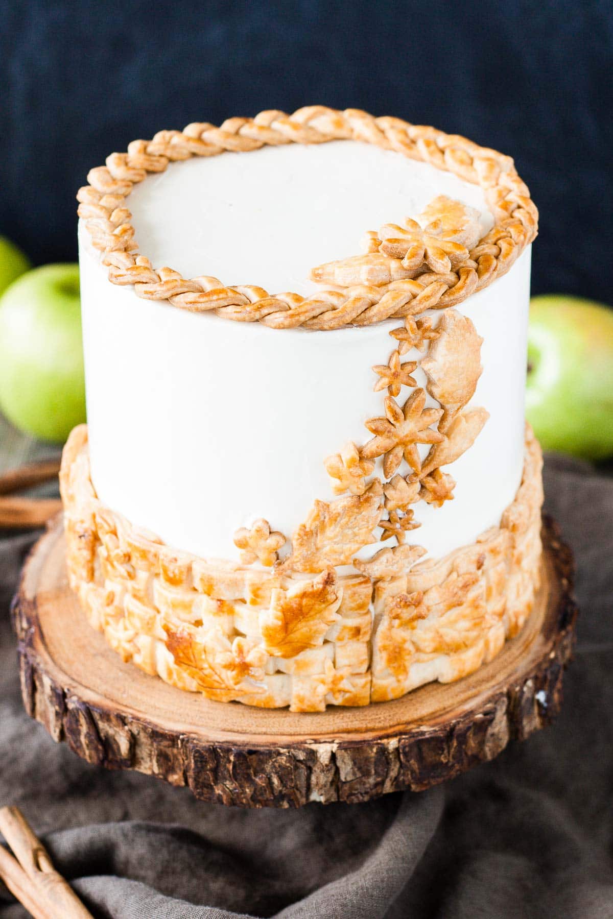 A cake sitting on top of a rustic wooden cake stand with apples in the background.