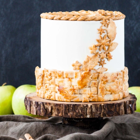 A cake sitting on top of a rustic wooden cake stand with apples in the background.