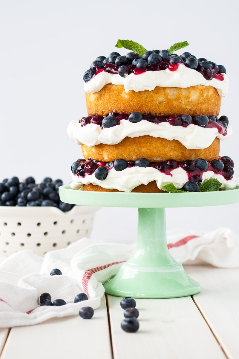 Blueberry shortcake decorated in a rustic, naked cake style on a green cake stand with a bowl of blueberries to the side.