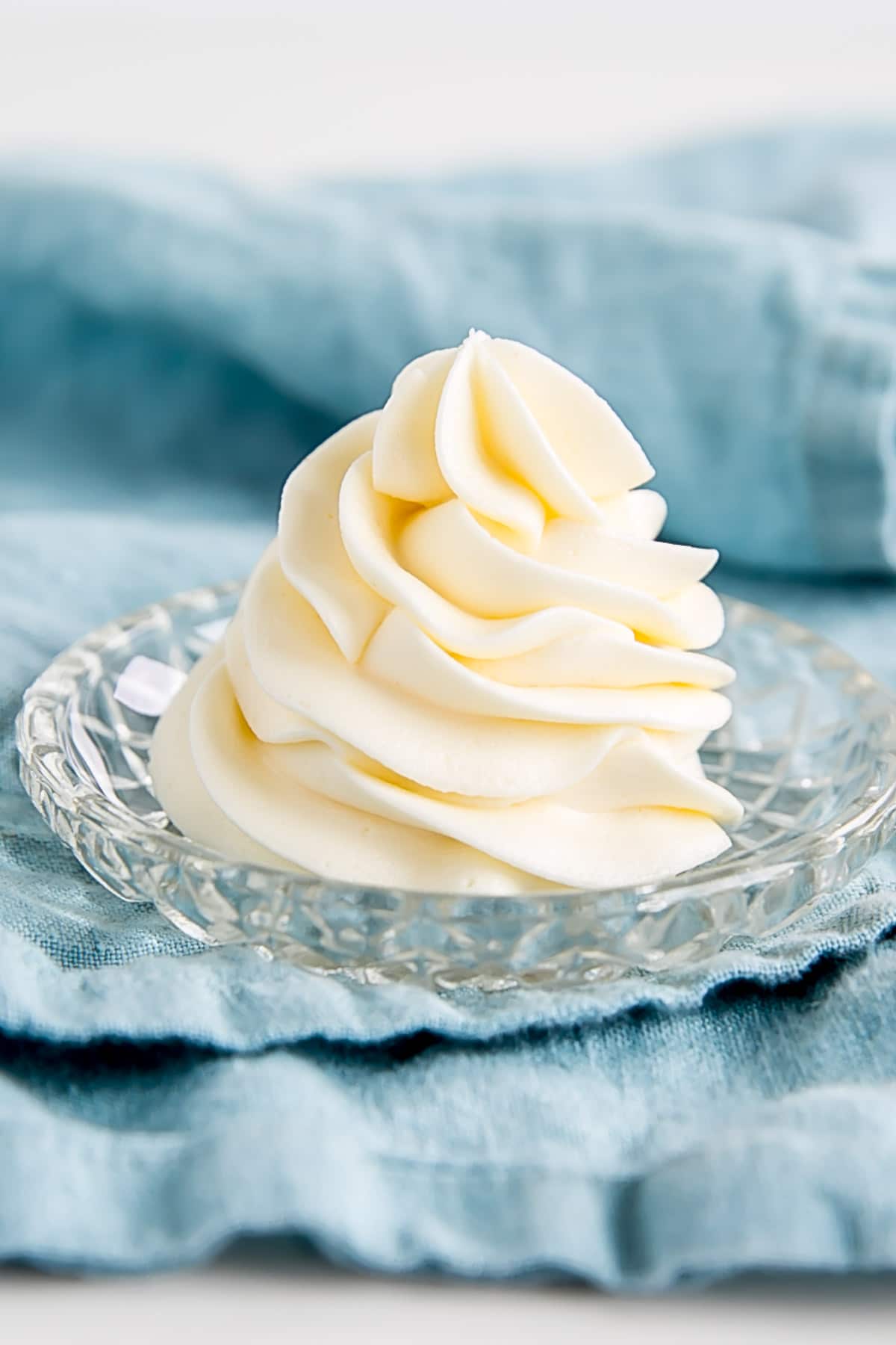 Piped swirl of Ermine Frosting on a small plate on a blue tea towel.