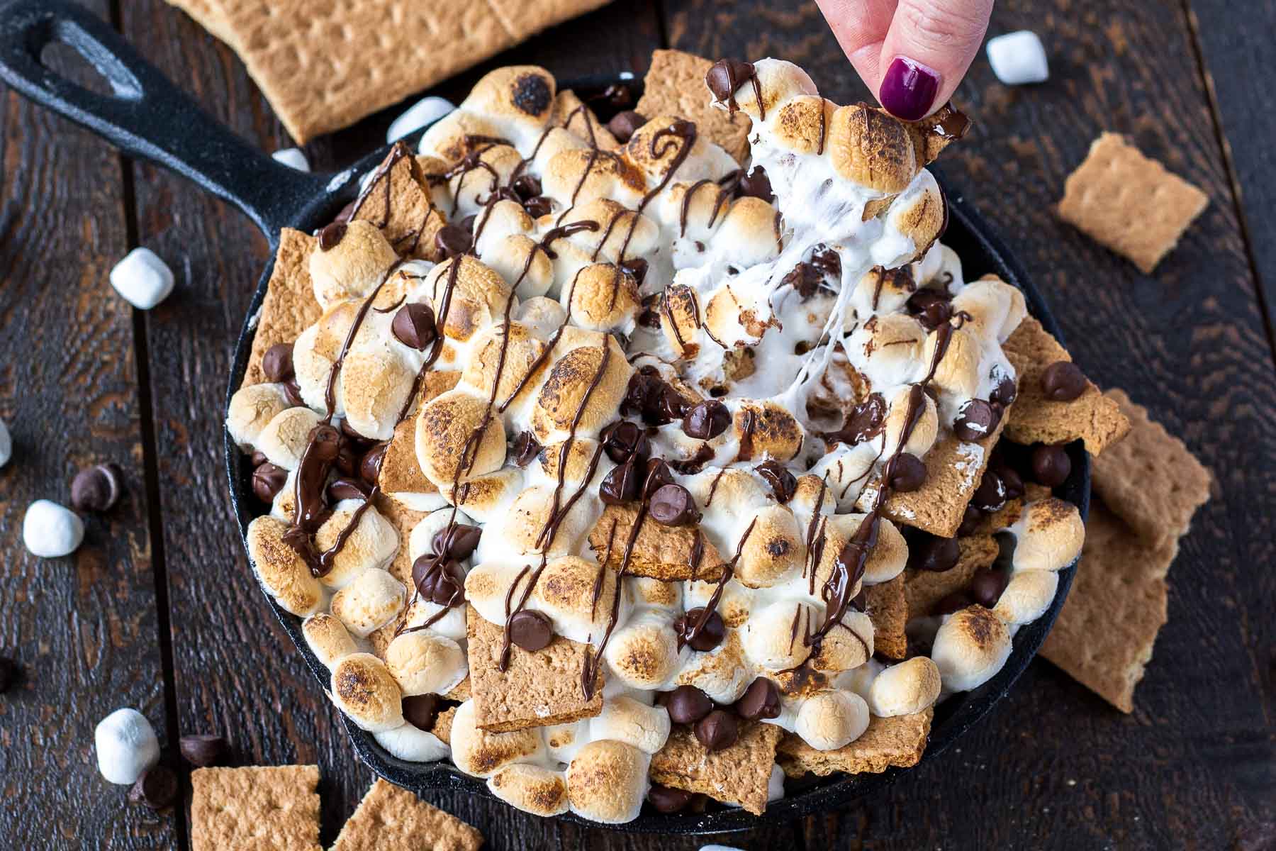 S\'mores nachos in a cast iron skillet. Fingers pulling out one of the \"nachos\".